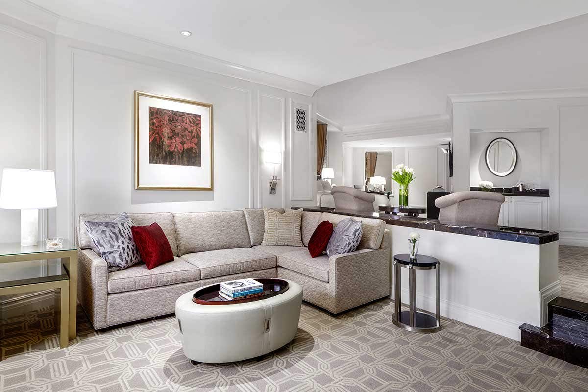 Hotel room with a couch, coffee table, and side table with a lamp. A king bed and two armchairs visible behind the couch on an elevated floor.