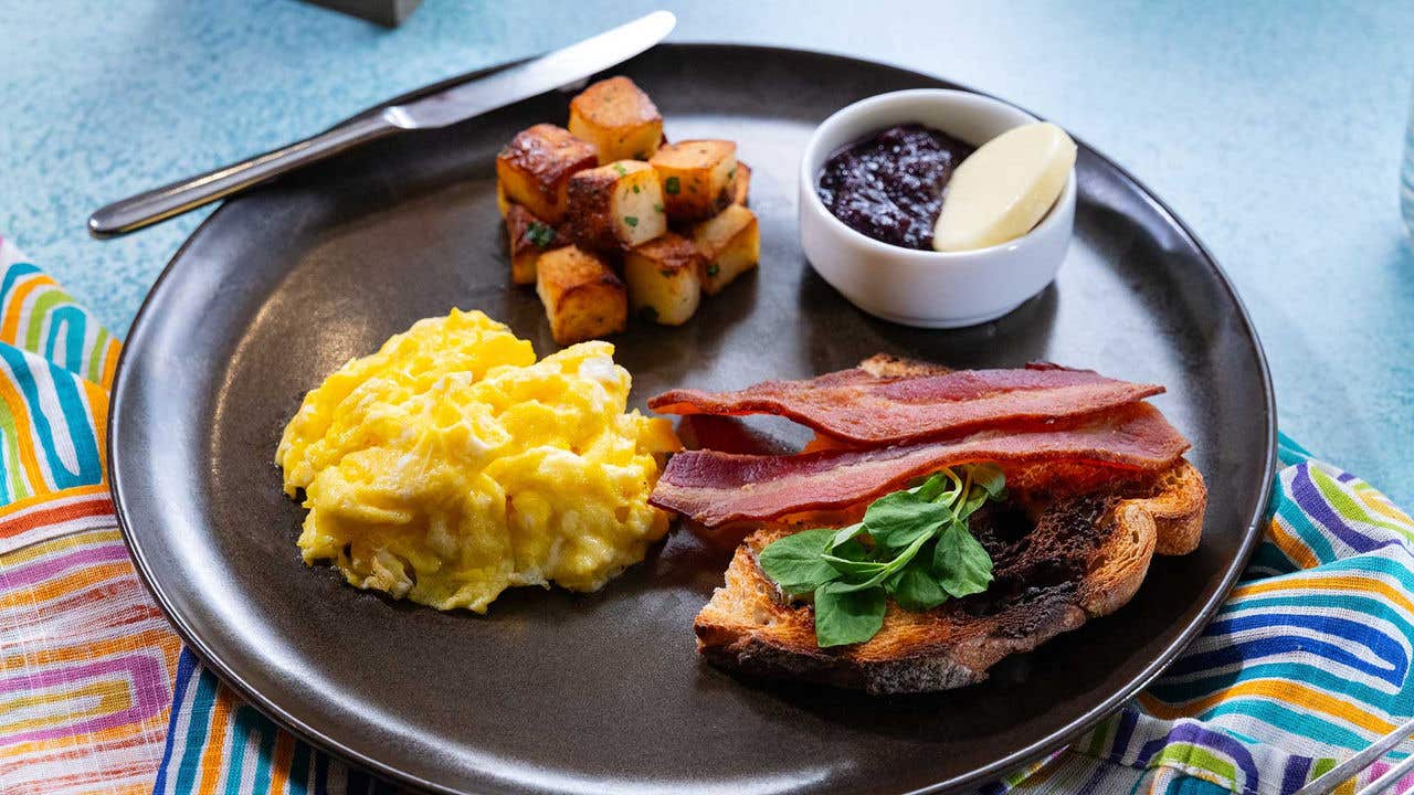 Scrambled eggs, bacon on toast, roasted potatoes, and jam served on a dark plate.