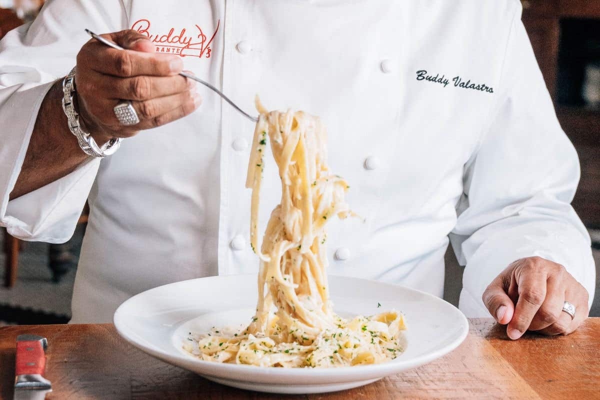 Close-up of a chef in a white jacket twirling fettuccine Alfredo on a fork above a plate of pasta.